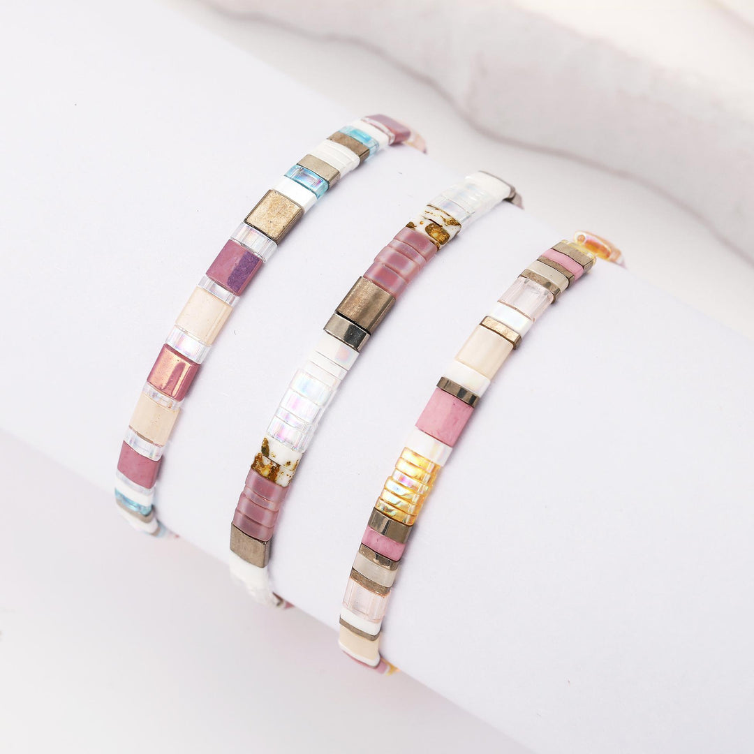 Three beaded stretch bracelets with rectangular glass beads in pink, gold, white, and blue, displayed on a white wristband.