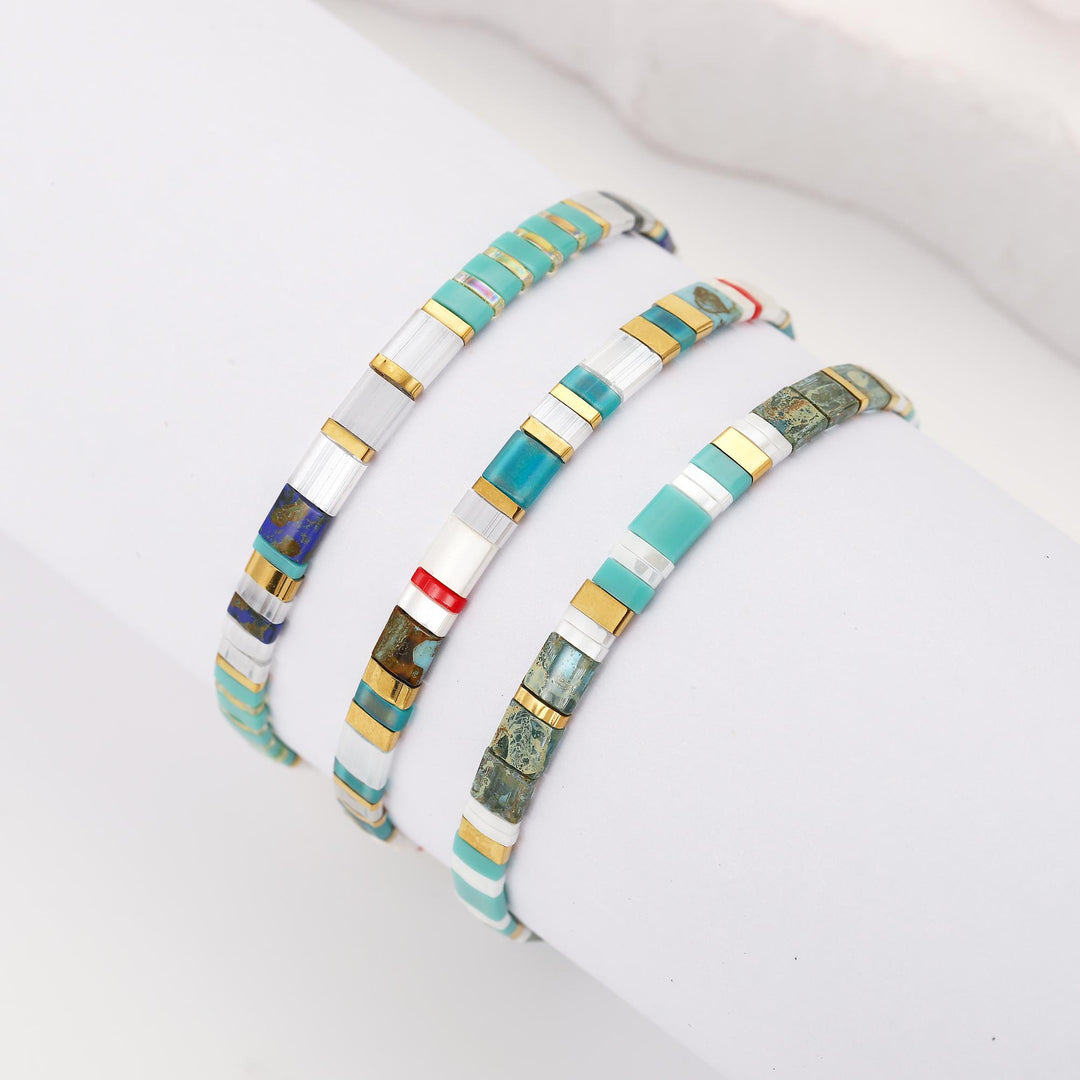 Three beaded bracelets displayed on a white cylindrical stand. The bracelets feature segments of turquoise, white shell, gold, and green stone, with one bracelet showing a small red accent bead.