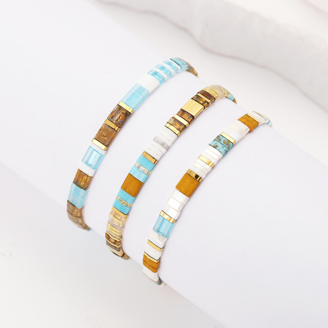 Three beaded stretch bracelets displayed on a white wrist form. The bracelets feature alternating segments of light blue, white, and metallic gold with some segments showing a textured brown pattern.
