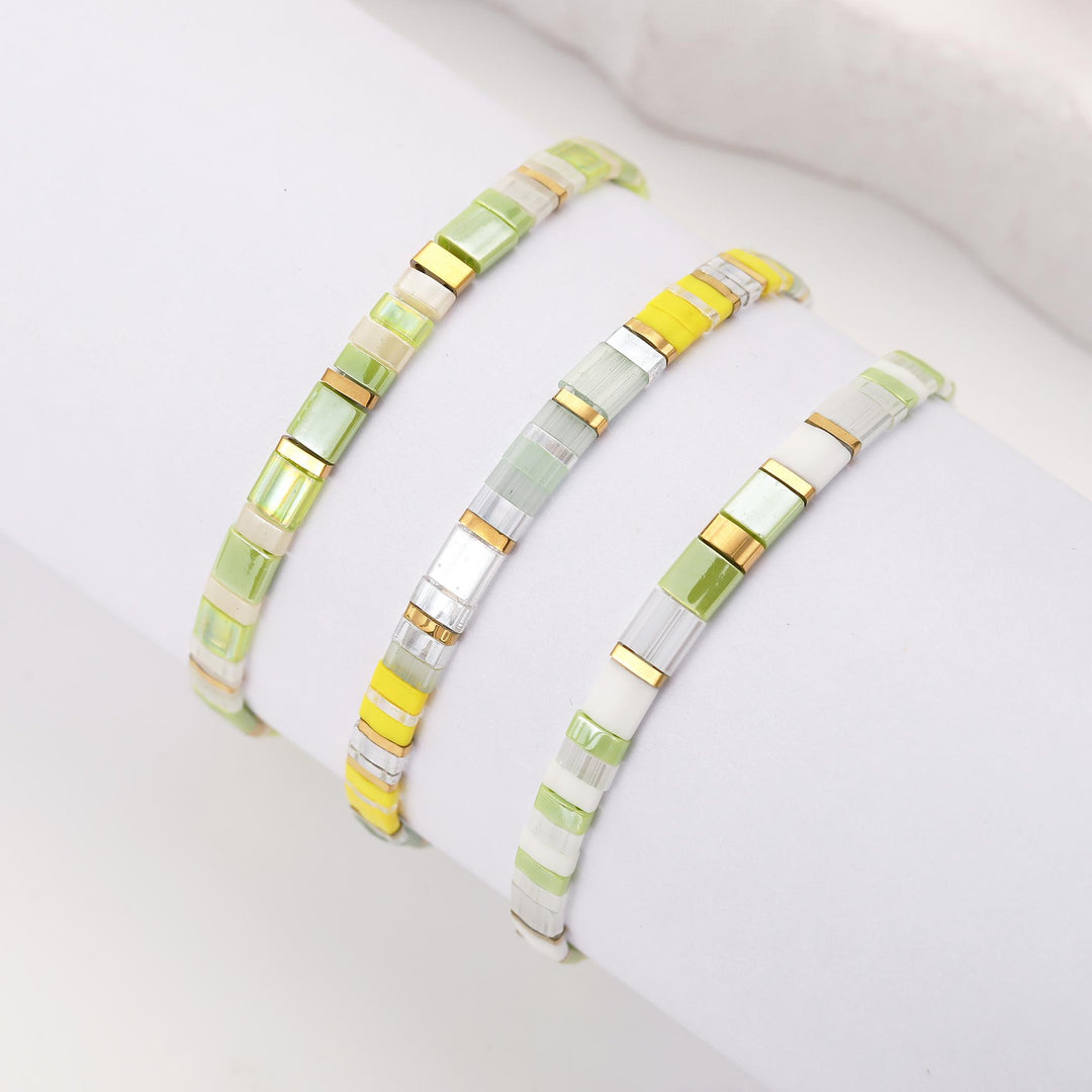 Three slim bangle bracelets with rectangular translucent beads in shades of light green, yellow, white, and clear, accented with metallic gold and silver, displayed on a white wrist form.