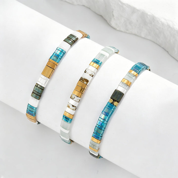 Three beaded stretch bracelets displayed on a white cylindrical stand. The bracelets feature segments of translucent blue, white, gold, and dark green, with one bracelet showing a small black bead accent.