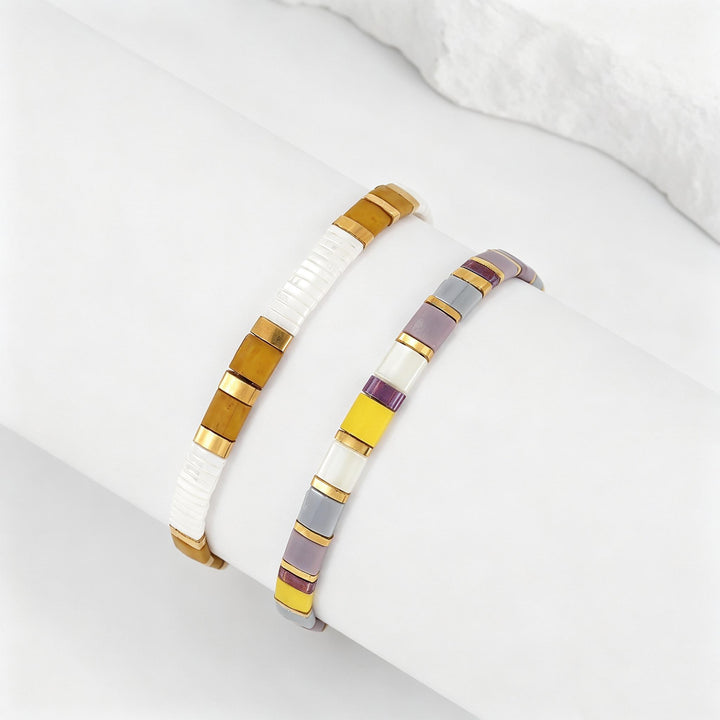 Two beaded stretch bracelets displayed on a white cylindrical form. One bracelet features white and gold beads, while the other features white, gray, yellow, and purple beads with gold accents.